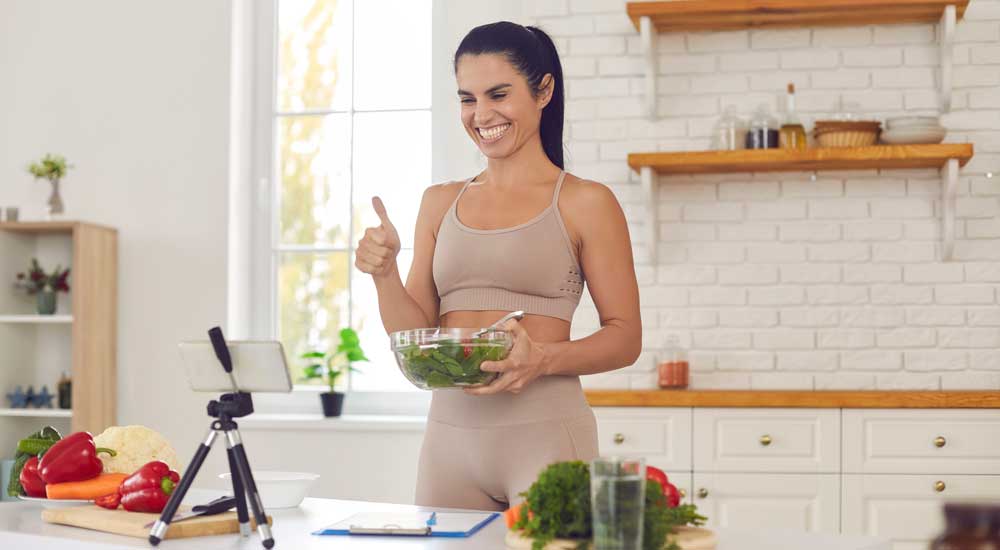 woman holds salad bowl with phone on tripod recording